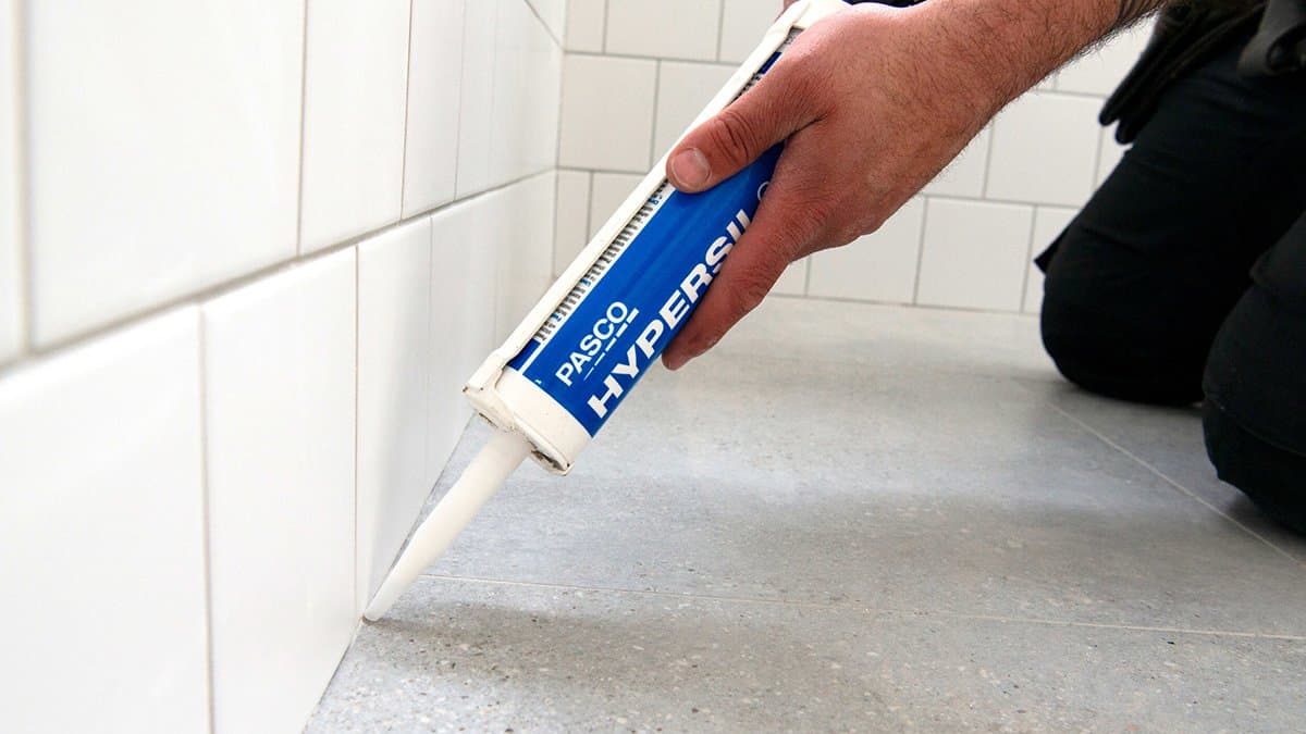 Hand applying a bead of silicone sealant along a tiled wall and floor junction