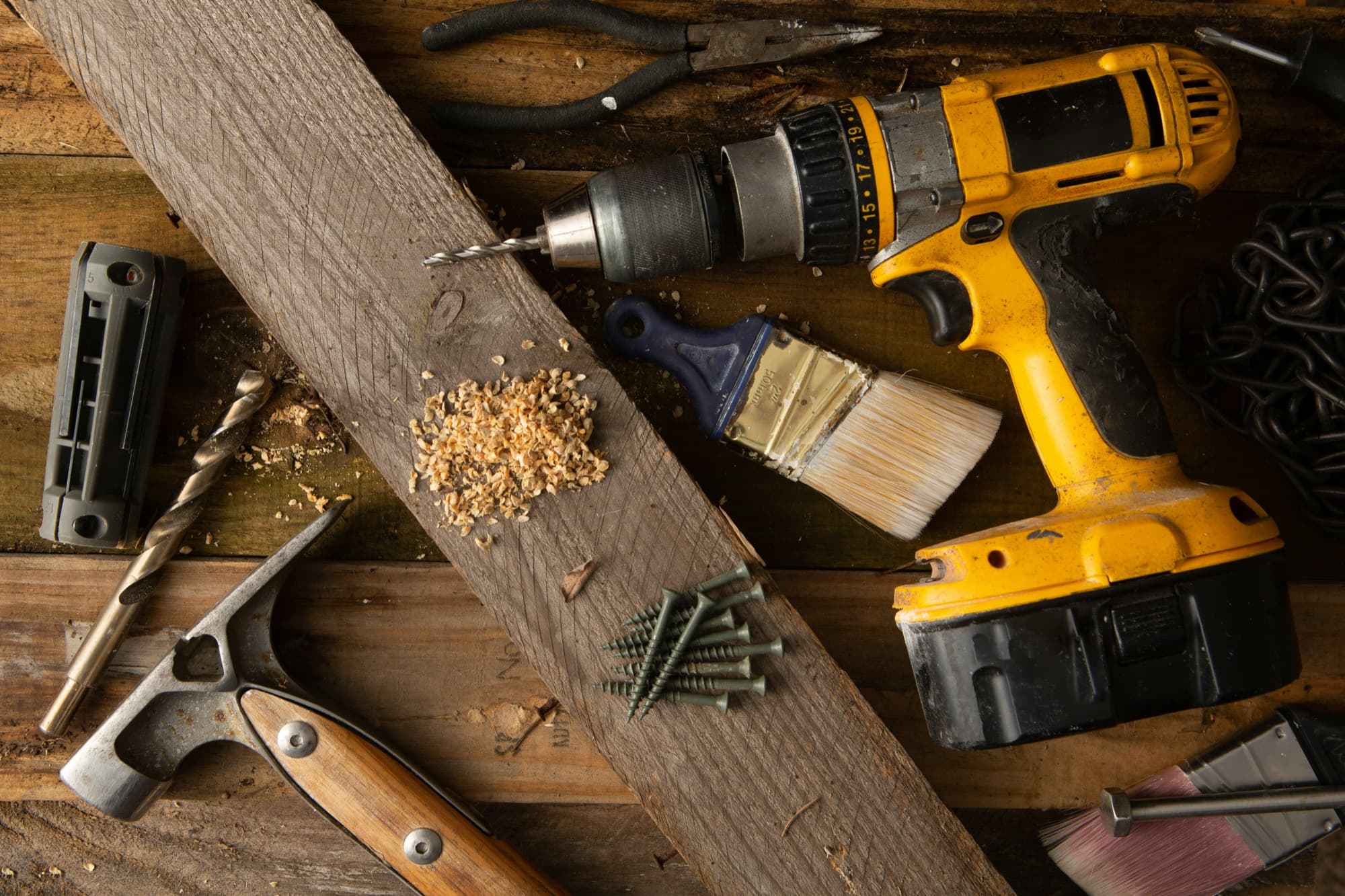 Overhead flatlay of a cordless drill, hammer, paintbrush and screws on a timber workbench