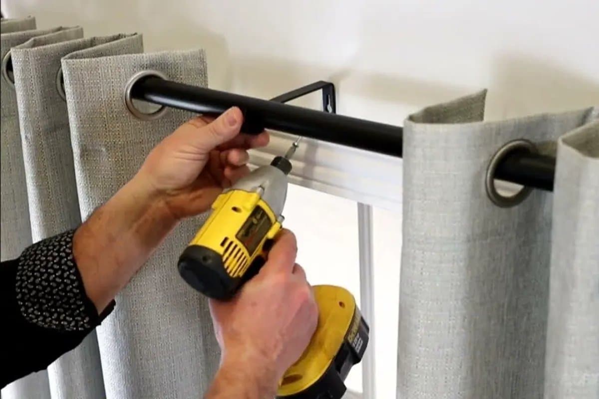 Hands using a cordless drill to fit a curtain rod bracket above a window
